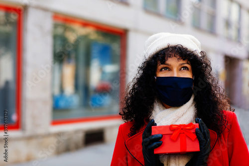 Portrait of a woman in red shopping outside with face mask holding christmas gift looking up © Carlos David