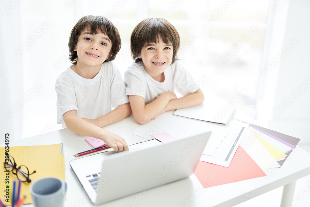 Best education for kids. Two adorable latin boys, brothers smiling at camera while sitting together at the table and using laptop. Little kids having online lesson at home Best education for kids. Two adorable latin boys, brothers smiling at camera while sitting together at the table and using laptop. Little kids having online lesson at home