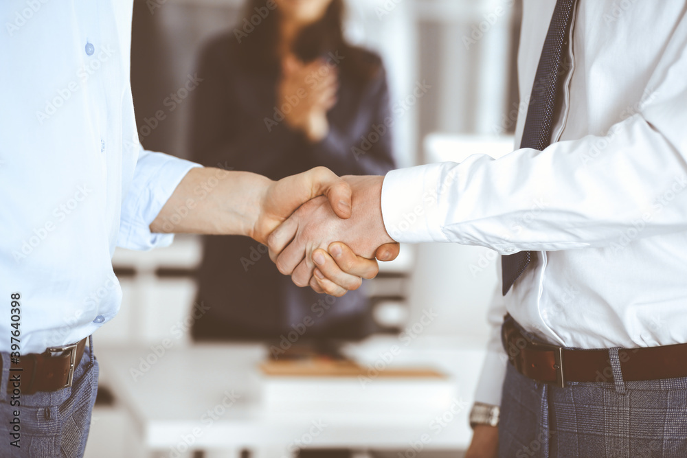 Fototapeta premium Two businessmen are shaking hands in office, close-up. Happy and excited business woman stands with raising hands at the background. Business people concept