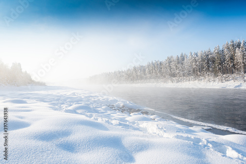 Frosty fog over winter river with snow and forest on bank. First ice on lake on cold day. © Koirill