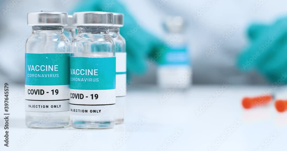 Doctor in protective uniform holding bottle with medication, coronavirus vaccine. Vaccine vials on a white laboratory table. Global vaccination. Doctor in protective uniform holding bottle with medication, coronavirus vaccine. Vaccine vials on a white laboratory table. Global vaccination.