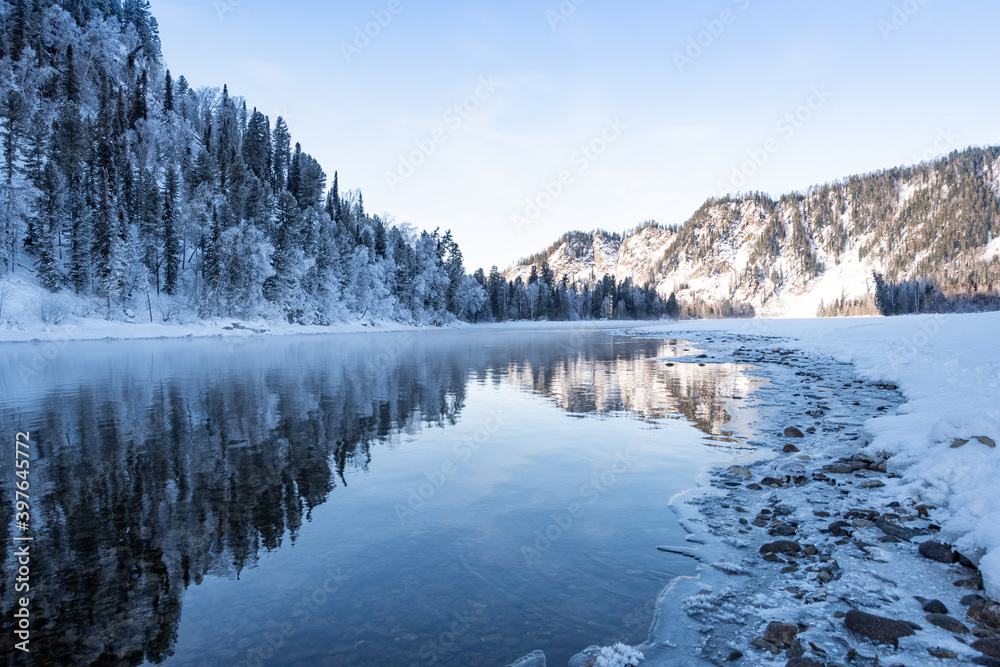 Snow forest on Bank of winter river. Reflection of frost trees in water.