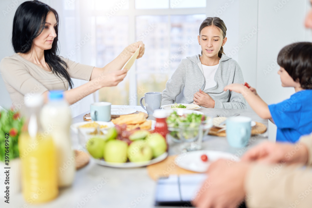 Pretty latin teenaged girl looking cheerful while having breakfast with her mom and little brother at home