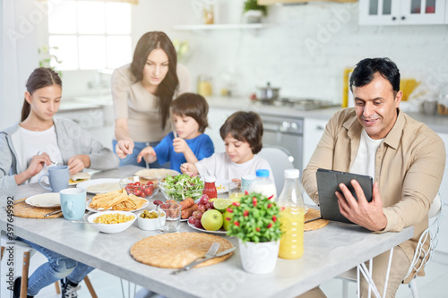Family routine. Smiling hispanic father using digital tablet while having lunch together with his family, sitting at the kitchen table at home