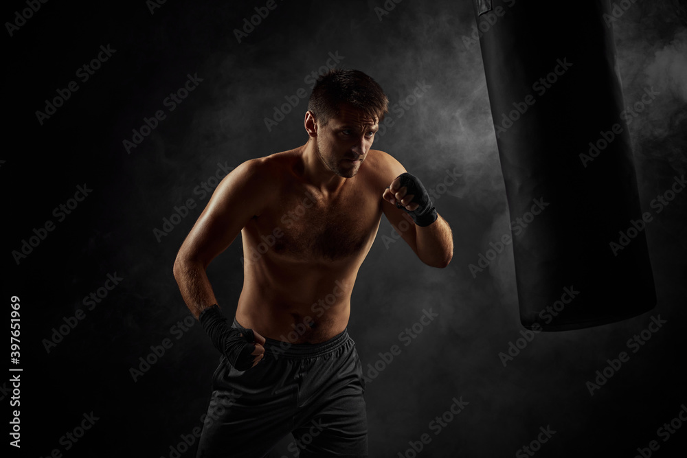 Aggressive shirtless boxer training defense and attacks in boxing bag on black background