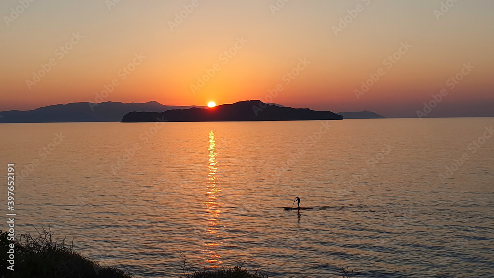 A man is sailing on a board with an oar during a beautiful golden sunset over the water on Crete, Greece.