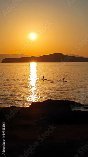A man is sailing on a board with an oar during a beautiful golden sunset over the water on Crete, Greece.