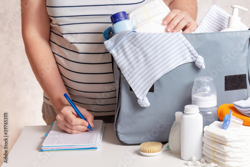 Unrecognizible pregnant caucasian woman in striped t-shirt packing big blue diaper bag to maternity hospital. Diapers, nappy, hat, bottle and other necessary things for newborn baby.