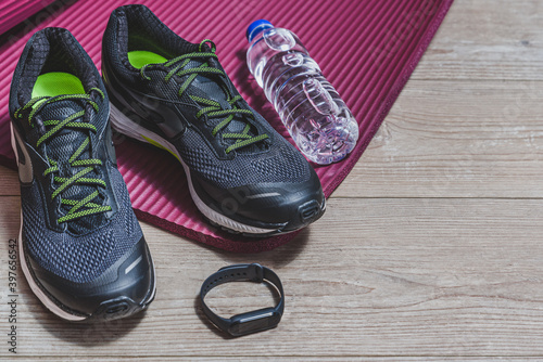 sport equipment with sport shoes, pilates mat, smart watch and bottle of water on a wooden floor