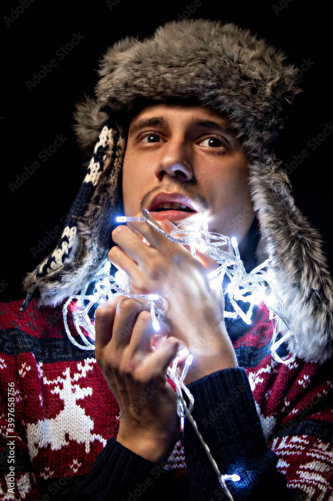 Fototapeta premium A young guy holds a garland in his hands. Christmas garland. The guy is holding a garland in front of him, a Christmas decoration. The light of the New Year's lights.