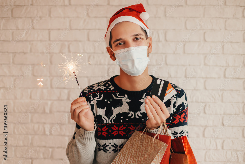Φωτογραφία Happy man shopper in Santa hat, medical face mask hold shopping bags, credit cards, sparkler light in hands