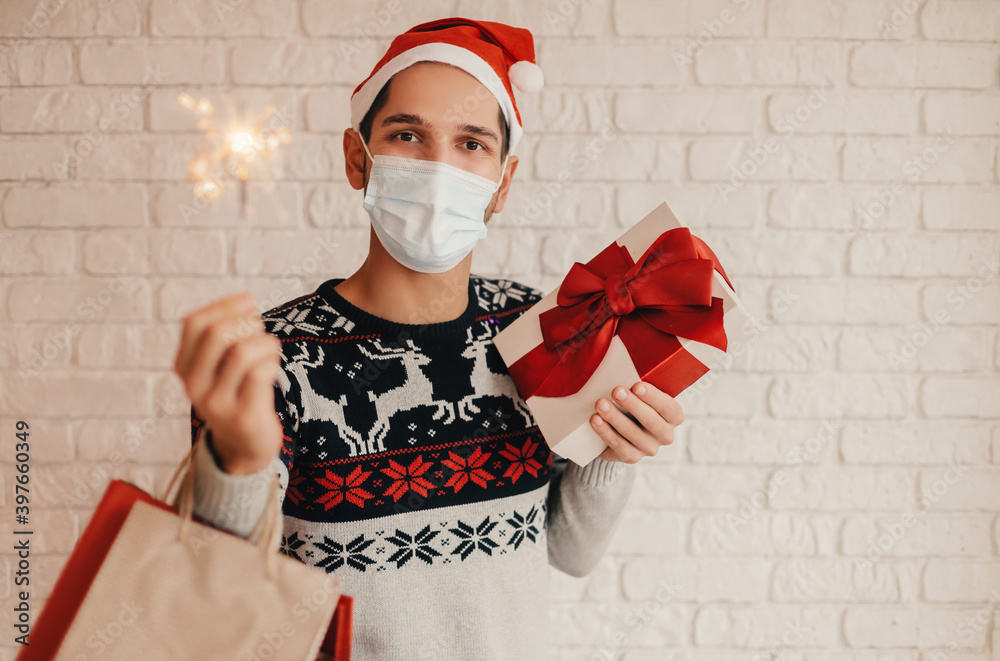 Φωτογραφία Happy man shopper in Christmas sweater, medical face mask hold shopping bag, gift box, sparkler light in hands