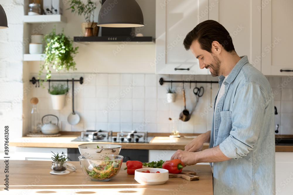 Side view smiling man cutting fresh vegetables, cooking homemade salad, standing at wooden table countertop in modern kitchen at home, preparing food for dinner or party, enjoying leisure time Side view smiling man cutting fresh vegetables, cooking homemade salad, standing at wooden table countertop in modern kitchen at home, preparing food for dinner or party, enjoying leisure time