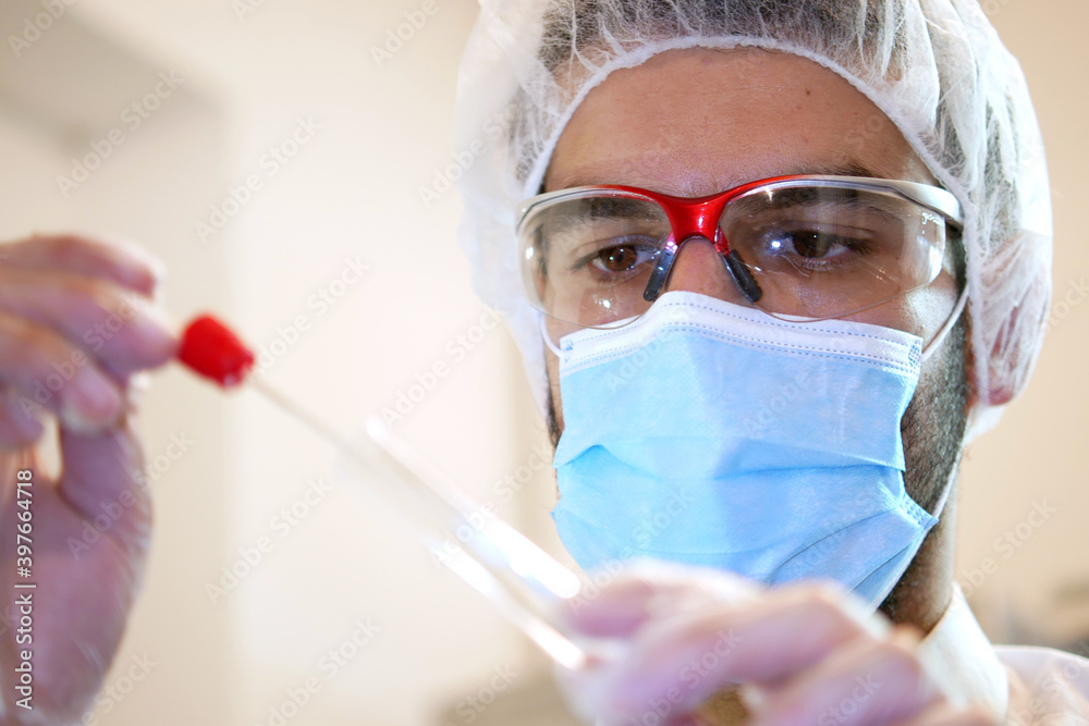Young attractive caucasian man scientist researching in the hospital laboratory, looks at the sample of throat and nose swab, close up of doctors face and hand with test tube 