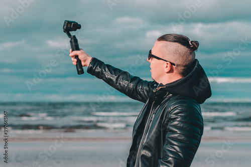 Young Blogger in Sunglasses Making Selfie or Streaming Video at the Beach Using Action Camera with Gimbal Camera Stabilizer. Hipster in Black Clothes Making Photo Against the Sea photo