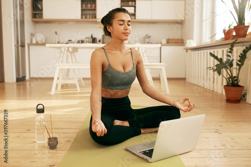 Attractive young Arabic female in top and leggings sitting in stylish home interior with open laptop, bottle of water and smoke of burning incense, practicing meditation, having calm peaceful look