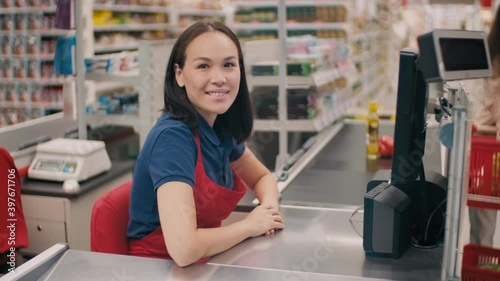 Medium portrait of friendly woman in uniform working as cashier in big supermarket smiling at camera while unrecognizable customer in mask placing products on conveyor belt in blurred background photo