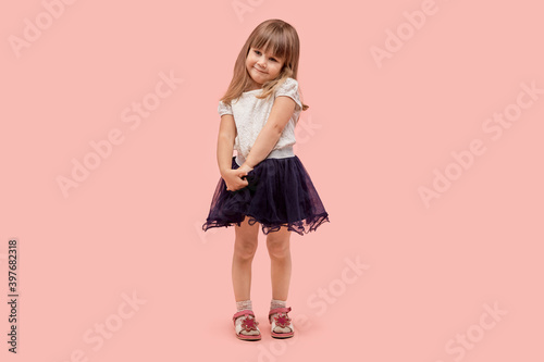 A cute, demure little girl in a black skirt and white tank top looks at the camera. Pink background.