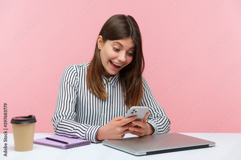 Excited shocked woman office worker surprised with sale and discounts doing shopping online on her smartphone sitting at workplace. Indoor studio shot isolated on pink background Excited shocked woman office worker surprised with sale and discounts doing shopping online on her smartphone sitting at workplace. Indoor studio shot isolated on pink background