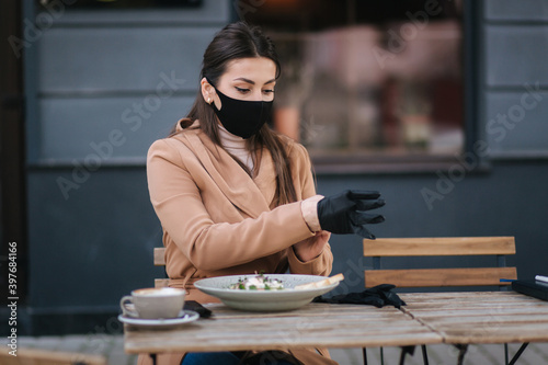 Yiung woman in black protective mask put on black gloves for having lunch. Female in protective mask sitting outdoors in cafe during quarantine covid -19 photo