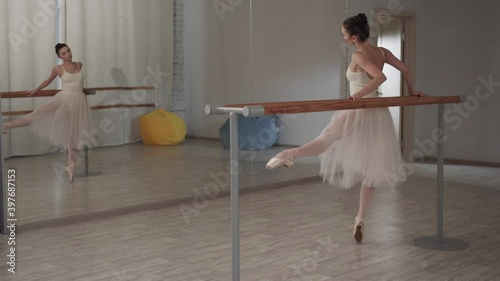 Lovely girl dancer in the ballet hall makes movements with her feet to the side and forward, standing in front of the mirror photo