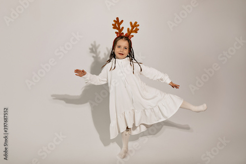 Full-lenght studio shot of little girl wearing white dress and Christmas accessories in the head dancing and having fun over isolated background