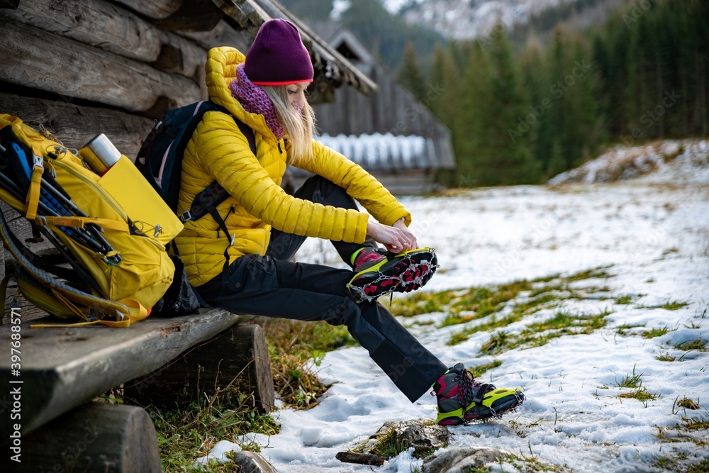 Woman hiker in a yellow down jacket puts yellow crampons on her shoes before going to the mountains. The backpack with equipment is lying next to it.