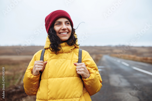 Portrait of a female tourist with a backpack wearing yellow jacket and red hat stands on the road. Young woman travels during winter or late autumn season. Hitch-hiking, trip and travelling concept. photo