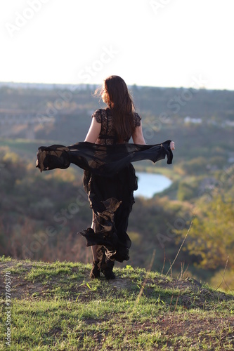 A beautiful girl stands on a cliff in a black dress. Nice view, river, nature. Black shawl.