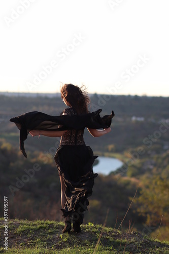 A beautiful girl stands on a cliff in a black dress. Nice view, river, nature. Black shawl.