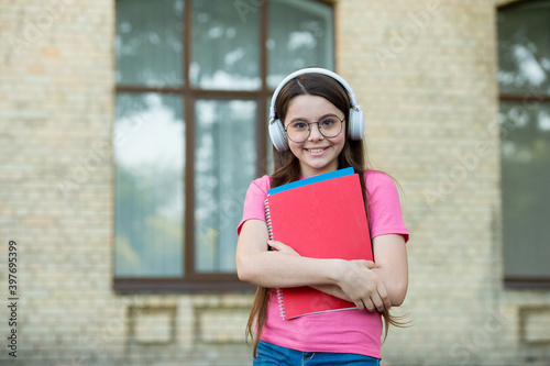 Happy kid wear modern headphones holding school books outdoors, audio course