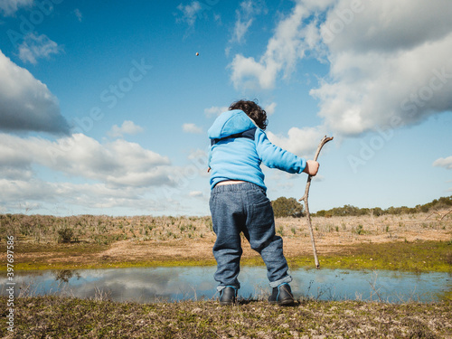 niño jugando a lanzar piedras al agua con abrigo bajo un cielo azul photo