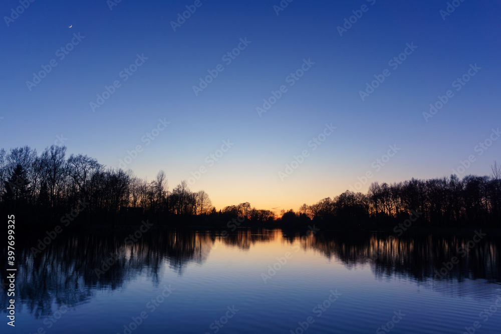 Clear blue sky after sunset at lake with reflecting silhouettes of trees