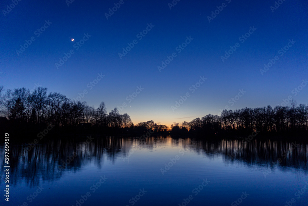 Clear blue sky after sunset at lake with reflecting silhouettes of trees, venus and crescent moon Clear blue sky after sunset at lake with reflecting silhouettes of trees, venus and crescent moon