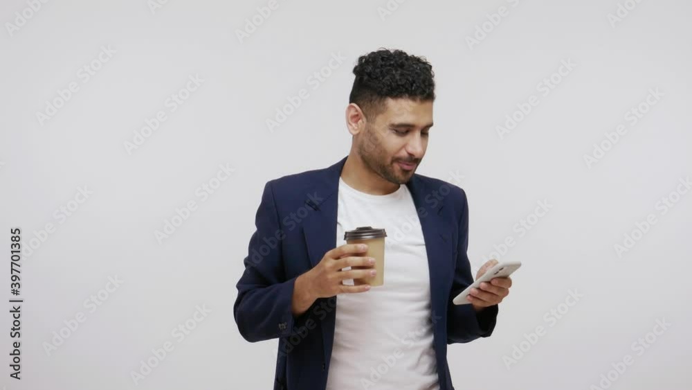 Busy man in elegant suit drinking coffee from paper cup and surfing on smartphone, booking tickets, buying goods, ordering delivery. Indoor studio shot isolated on gray background Busy man in elegant suit drinking coffee from paper cup and surfing on smartphone, booking tickets, buying goods, ordering delivery. Indoor studio shot isolated on gray background