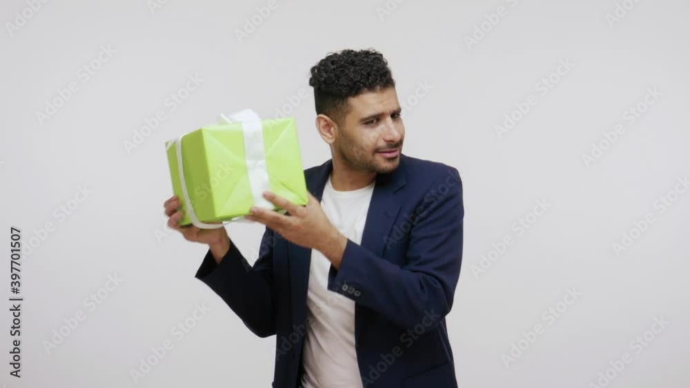 Curious bearded man in elegant suit shaking big green giftbox, trying to guess what is inside, untying bow. Indoor studio shot isolated on gray background Curious bearded man in elegant suit shaking big green giftbox, trying to guess what is inside, untying bow. Indoor studio shot isolated on gray background