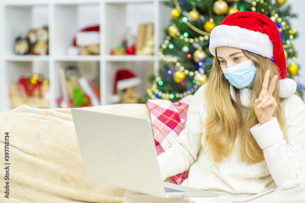 A girl in a santa hat and a medical mask waves a greeting to a laptop and congratulates online friends or relatives with merry christmas or new year during lockdown A girl in a santa hat and a medical mask waves a greeting to a laptop and congratulates online friends or relatives with merry christmas or new year during lockdown