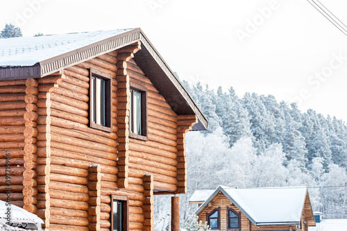 Close-up of a wooden house , the roof is covered with snow, smoke comes out of the chimney , in the background a winter forest and a small village