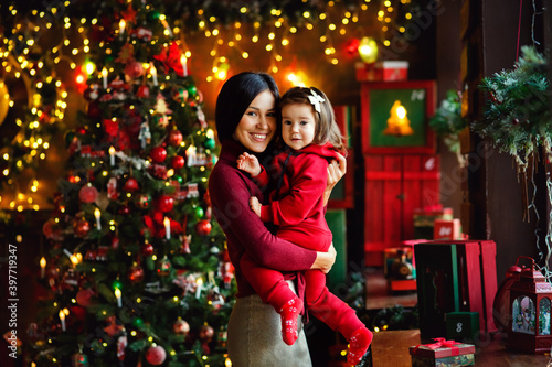 The Christmas family. Red background of a merry Christmas tree. A woman-mother and a child-daughter celebrate the New year, look at the camera together. Christmas greeting card.