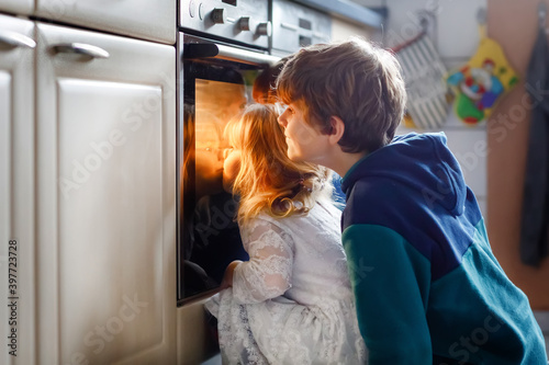 Little toddler girl and two kids boys baking, pizza or cookies in kitchen. Three children, siblings, brothers and sister sitting near oven and waiting. On thanksgiving or christmas holiday.