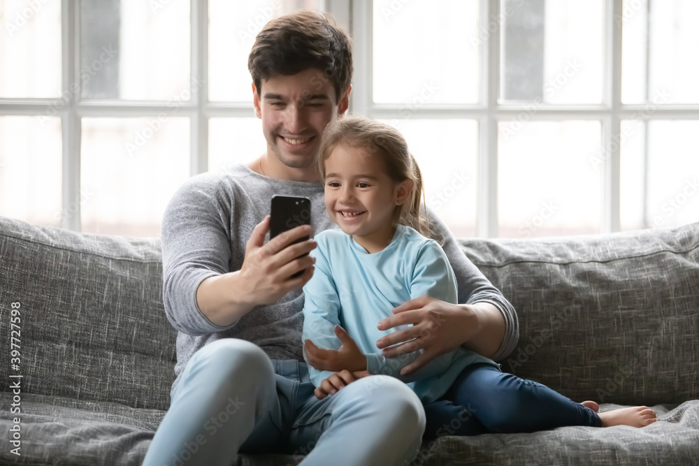 Smiling handsome young father embracing adorable small kid daughter, looking at smartphone web camera, positing for selfie photo, recording video or holding distant videocall, sitting on sofa.