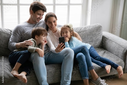 Relaxed young parents sitting on cozy sofa with cute little preschool kids siblings, using smartphone together, posing for selfie photo or recording funny video at home, modern technology concept.