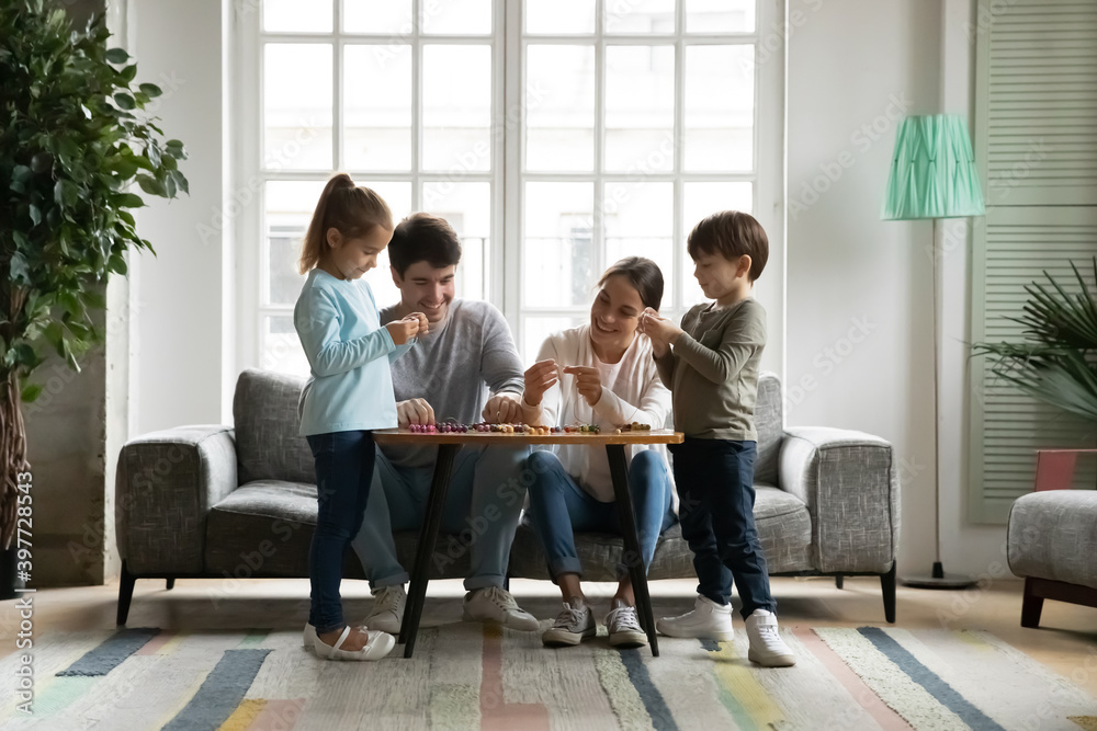 Full length smiling young affectionate parents sitting on sofa, involved in creative handicraft activity with interested little preschool kids siblings, making wooden accessories together at home. Full length smiling young affectionate parents sitting on sofa, involved in creative handicraft activity with interested little preschool kids siblings, making wooden accessories together at home.