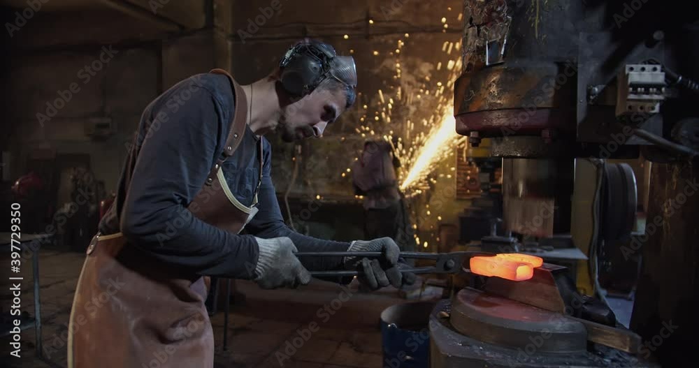 Two blacksmiths work in the smithy, one stands sideways to the camera and forges hot metal with a mechanical hammer, the second person stands with his back to the camera and cuts the metal with a saw Two blacksmiths work in the smithy, one stands sideways to the camera and forges hot metal with a mechanical hammer, the second person stands with his back to the camera and cuts the metal with a saw