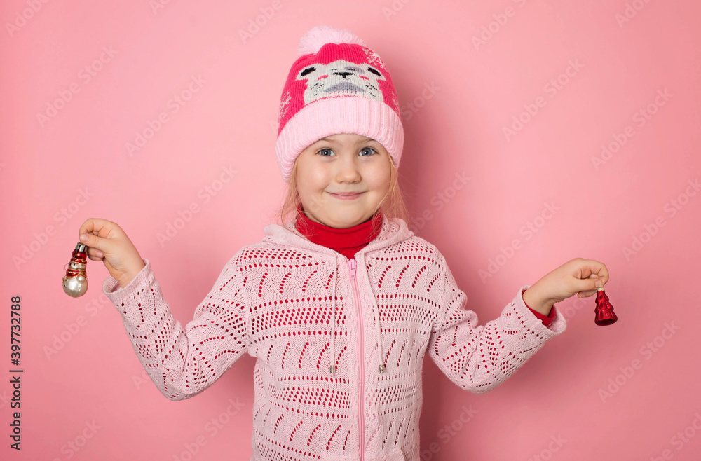 Smiling funny girl in pink winter hat holding Christmas decorations on pink background. Smiling funny girl in pink winter hat holding Christmas decorations on pink background.