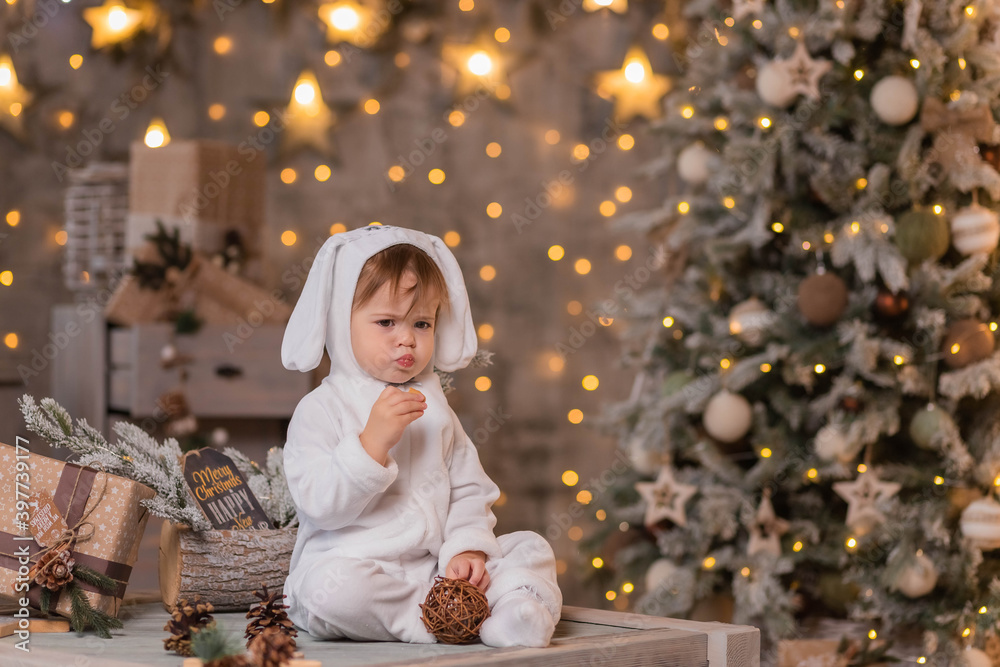 Merry Christmas and Happy New Year. A small child in a white rabbit costume laughs and sits against the background of a New Year's interior with a beautifully decorated Christmas tree. Merry Christmas and Happy New Year. A small child in a white rabbit costume laughs and sits against the background of a New Year's interior with a beautifully decorated Christmas tree.