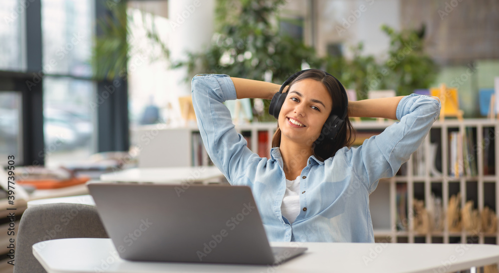 Fototapeta premium Beautiful happy young woman or student female employee sitting at table in the university library or office with laptop looking at the camera