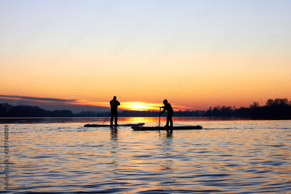 Silhouettes of two people rowing on supboards at sunset in winter