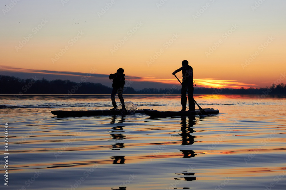 Silhouettes of two boys rowing on supboards (SUP) in winter river at sunset