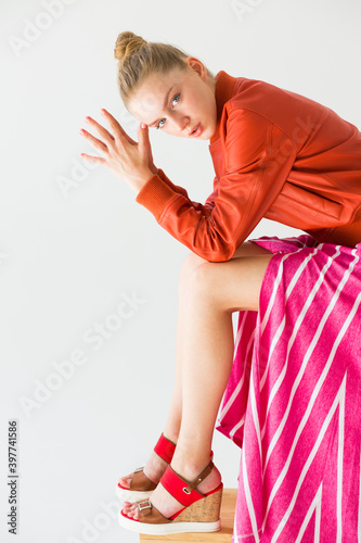The girl sits with her elbows leaning on her knees. The palms are folded and close to the head. Clothes - orange jacket, striped skirt and high wedge sandals.